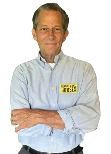Dennis with arms crossed in a blue button down featuring a “Dennis Buys Houses” logo, standing confidently on a transparent background.